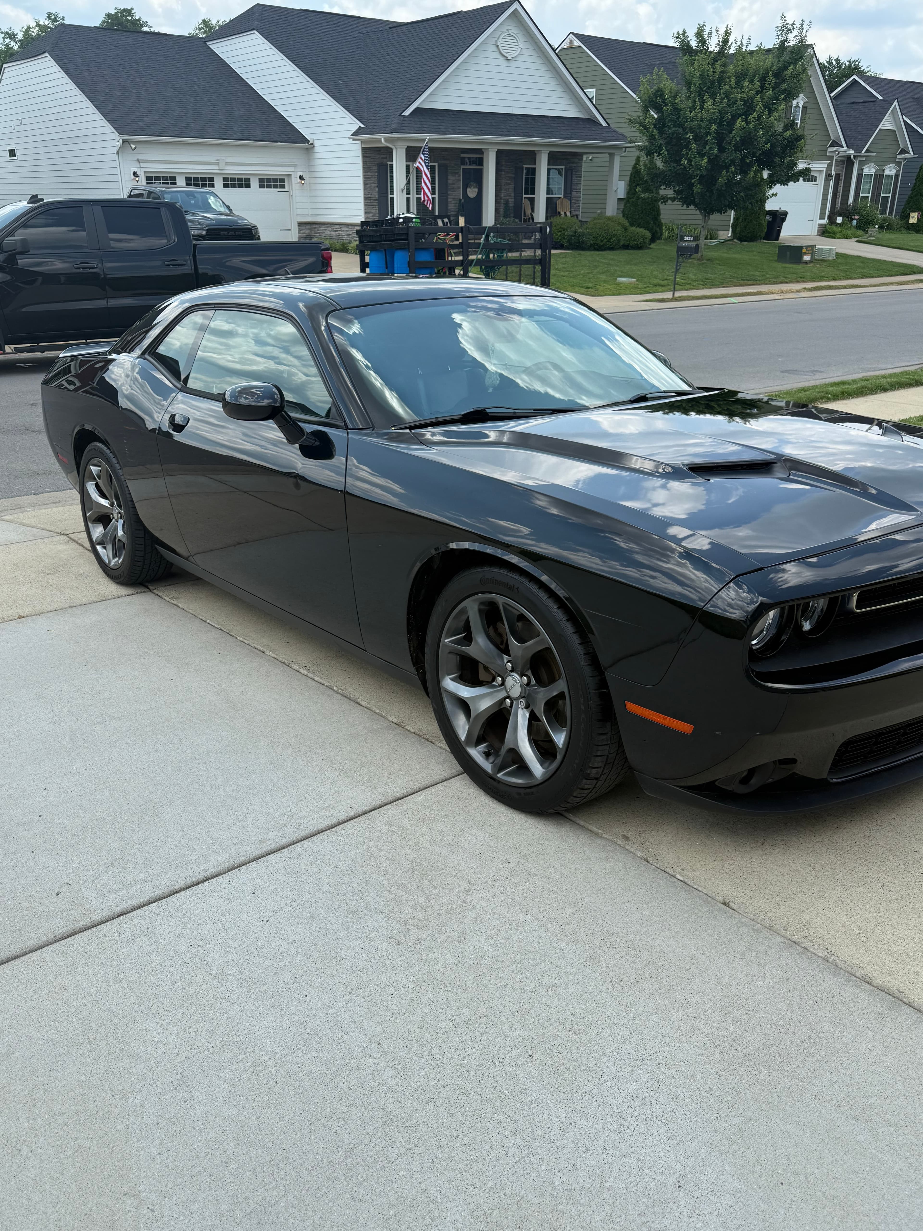 Shiny black Dodge Challenger parked on a concrete driveway reflecting clouds in a suburban neighborhood.