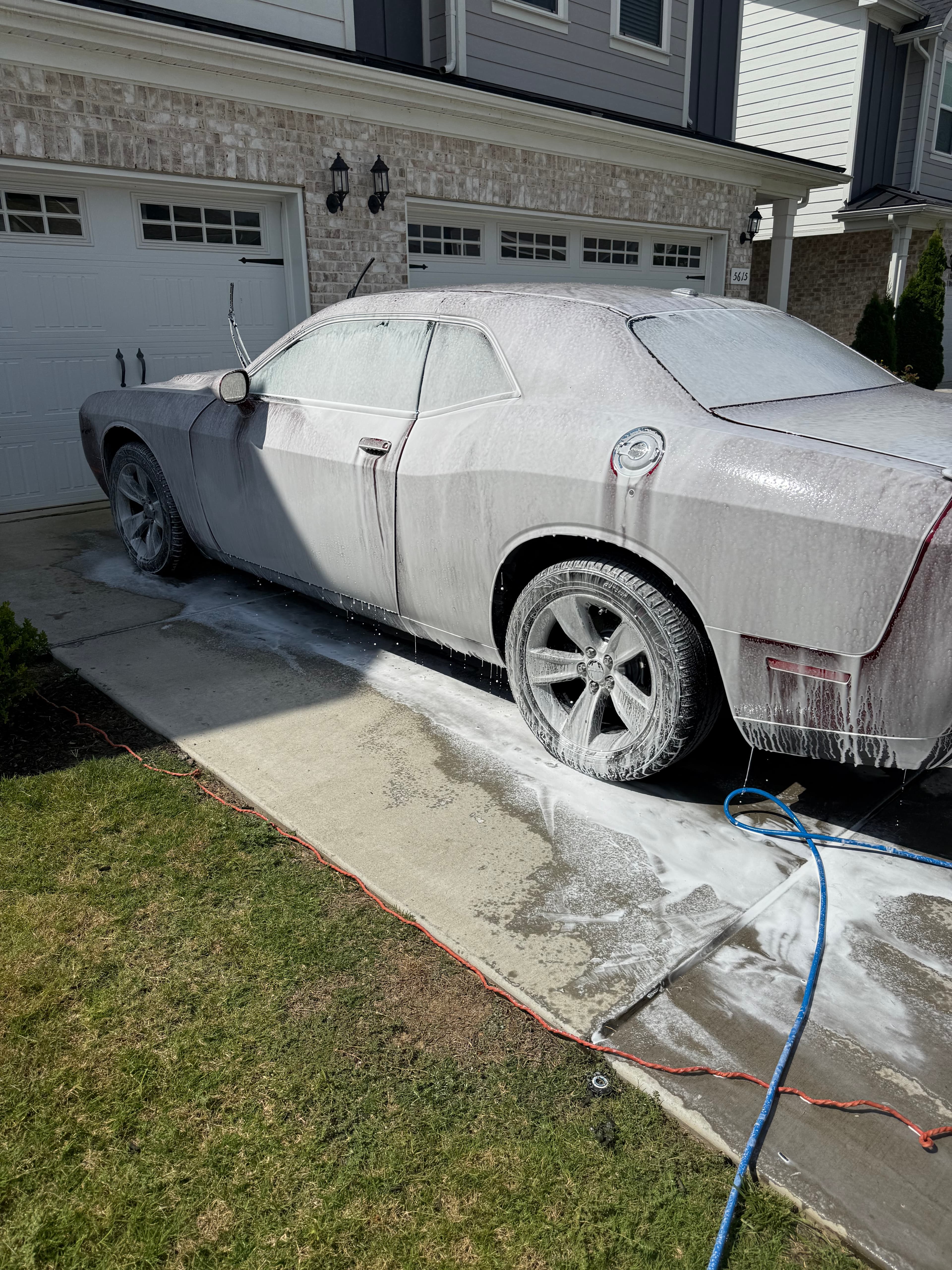 White Dodge Challenger covered in thick soap foam being washed on a residential driveway.
